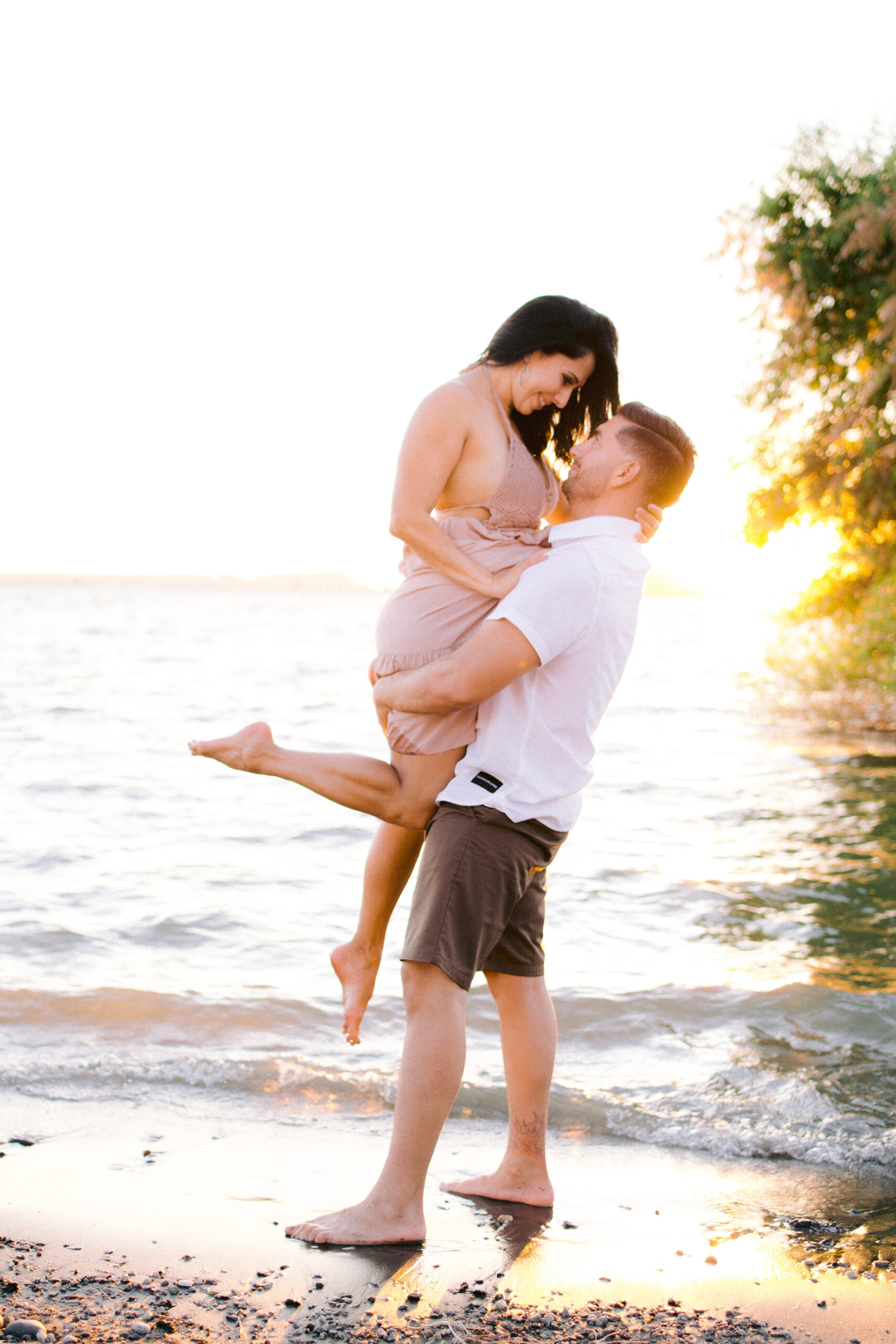 Man in white shirt and khaki shorts holding up woman in peach dress In front of lake during sunset.