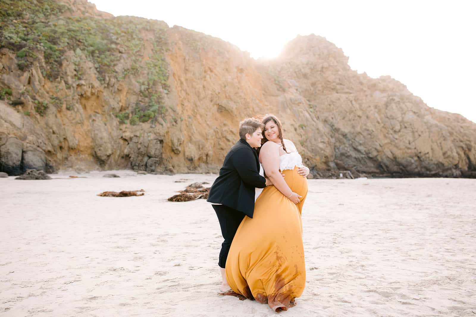 Lesbian Maternity photos in Big Sur on Beach. Two women hold each other on beach at sunset. One woman is pregnant