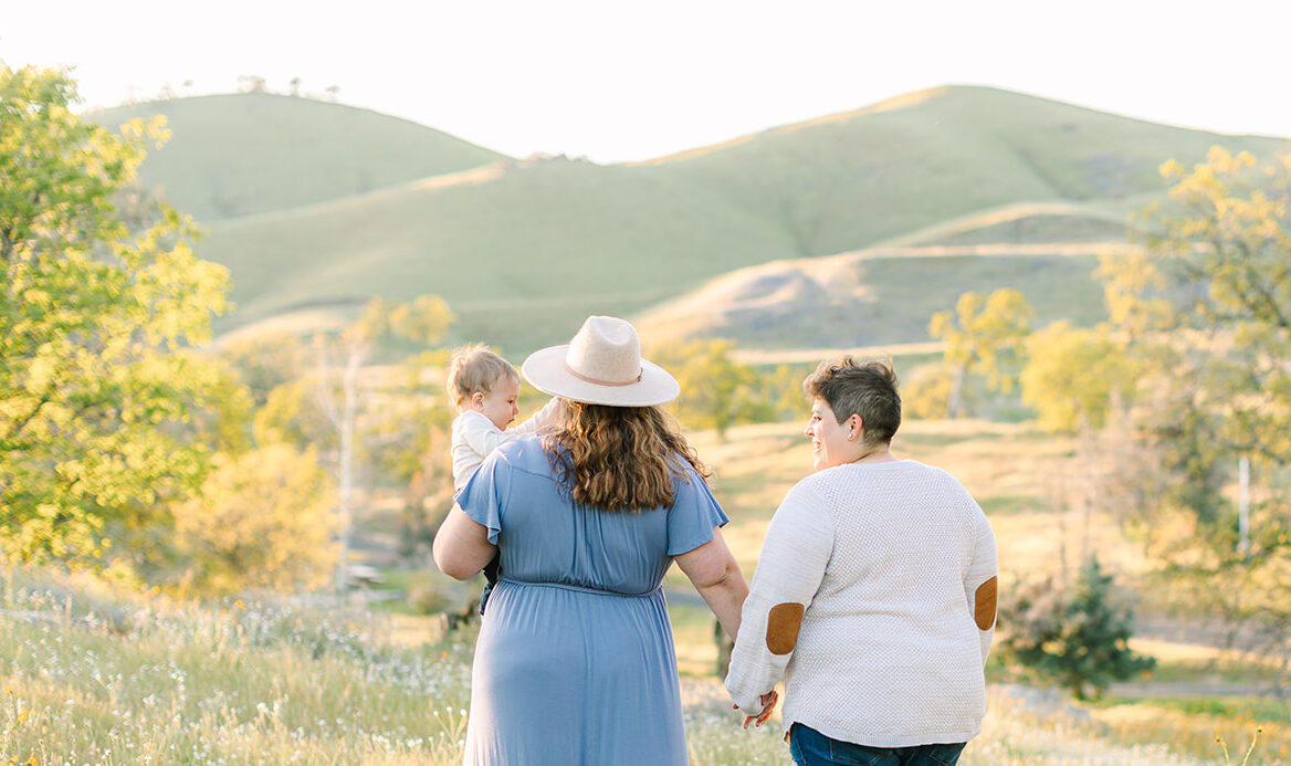 Family walking through golden grass in the Fresno foothills during a spring photoshoot, holding hands with their toddler in soft, natural light.