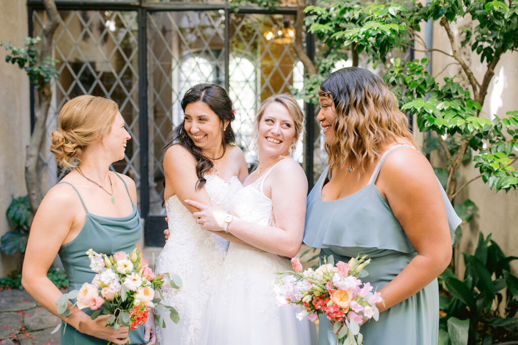 Brides embrace and smile with bridesmaids holding peach and pink bouquets at a Berkley City Club wedding, captured by Michelle Gunn Photo during a vibrant lesbian wedding celebration