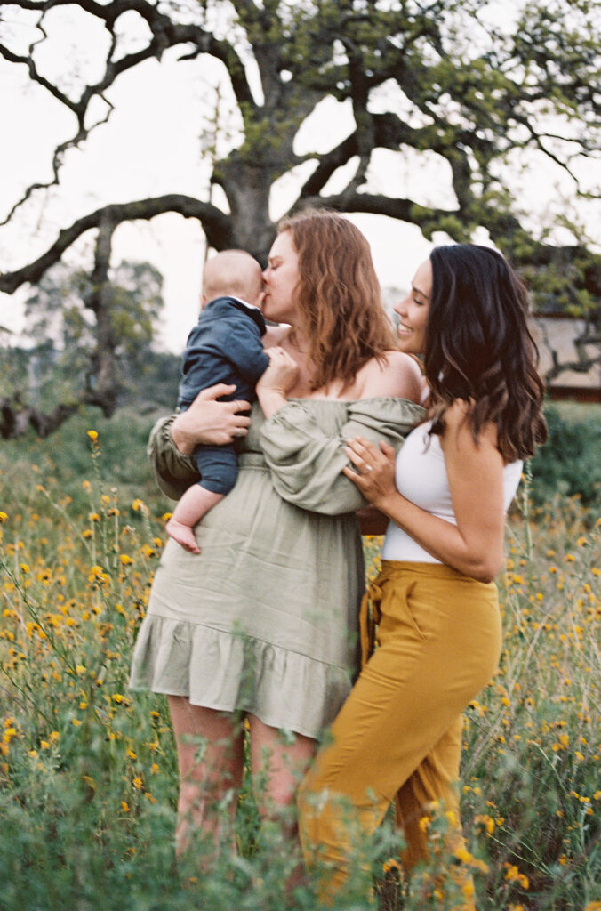 One mom kissing their baby’s head while the other mom smiles beside them under an oak tree, tender lesbian family photos captured on 35mm film.
