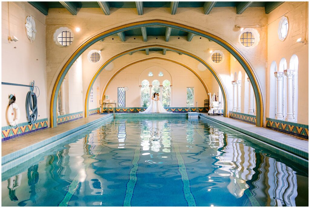 Brides stand together at the far end of the indoor pool during their Berkley City Club wedding, framed by arched architecture and reflected in the water at this lesbian wedding photographed by Michelle Gunn Photo.