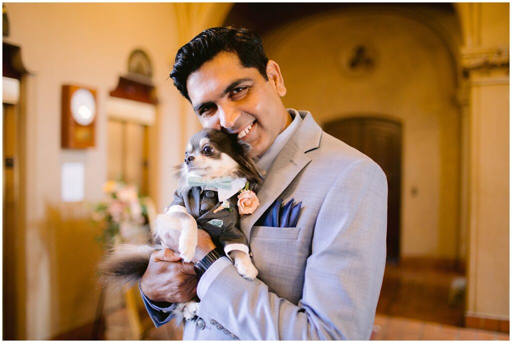 Man in a gray suit smiles while holding a chihuahua dressed in a tuxedo at a Berkley City Club wedding, captured by Michelle Gunn Photo during a romantic lesbian wedding celebration.