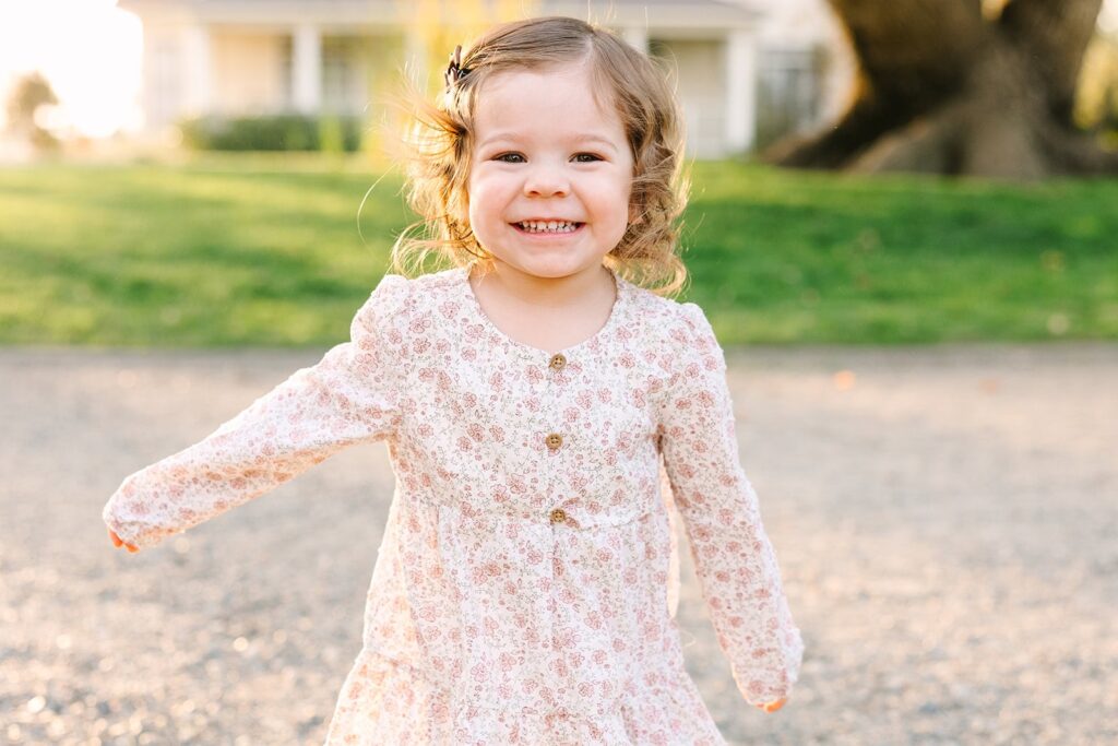Smiling toddler in a pink floral dress walking toward the camera in soft golden light during a Fresno family session, a joyful example of natural connection in Fresno family photo tips.