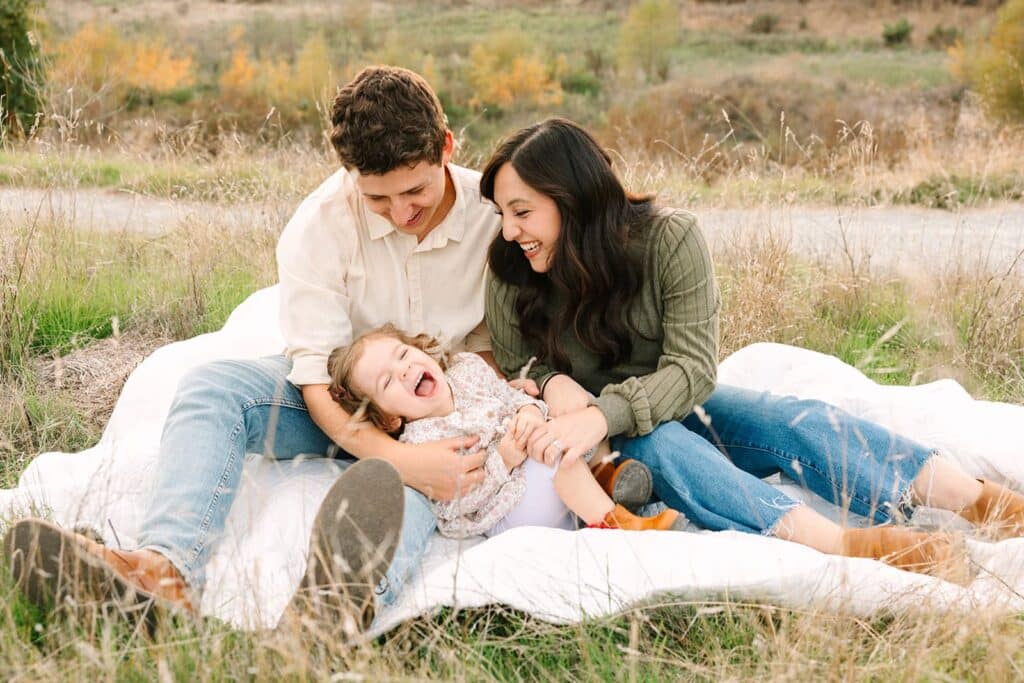 A family of three sitting on a blanket in a grassy field, laughing together during a Fresno family photo session, demonstrating playful posing ideas from Fresno family photo tips.