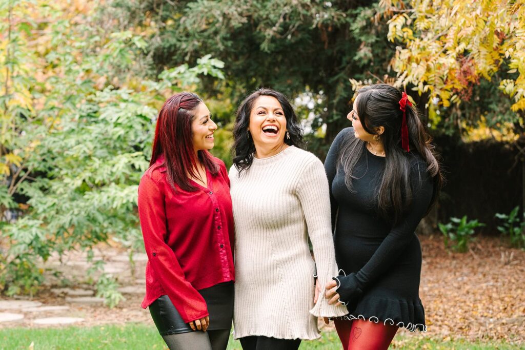 Three women standing together laughing in a leafy park during a Fresno family photo session, capturing natural interaction from Fresno family photo tips.