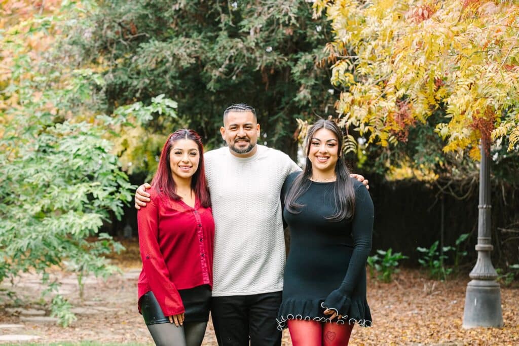Three family members standing with their arms around each other in a park with fall foliage during a Fresno family photo session, highlighting close connection from Fresno family photo tips.