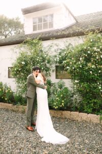 Full-body photo of bride and groom hugging in a flower-filled courtyard during their Fresno wedding