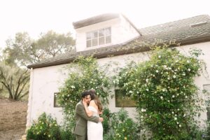 Bride and groom embracing in front of a white building covered in garden flowers during their Fresno wedding