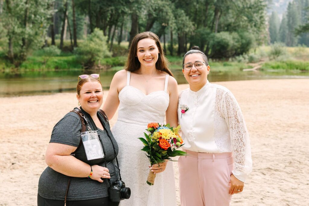 Photographer posing with a couple holding a bouquet on a sandy riverbank surrounded by trees, photographed by michelle gunn photo.